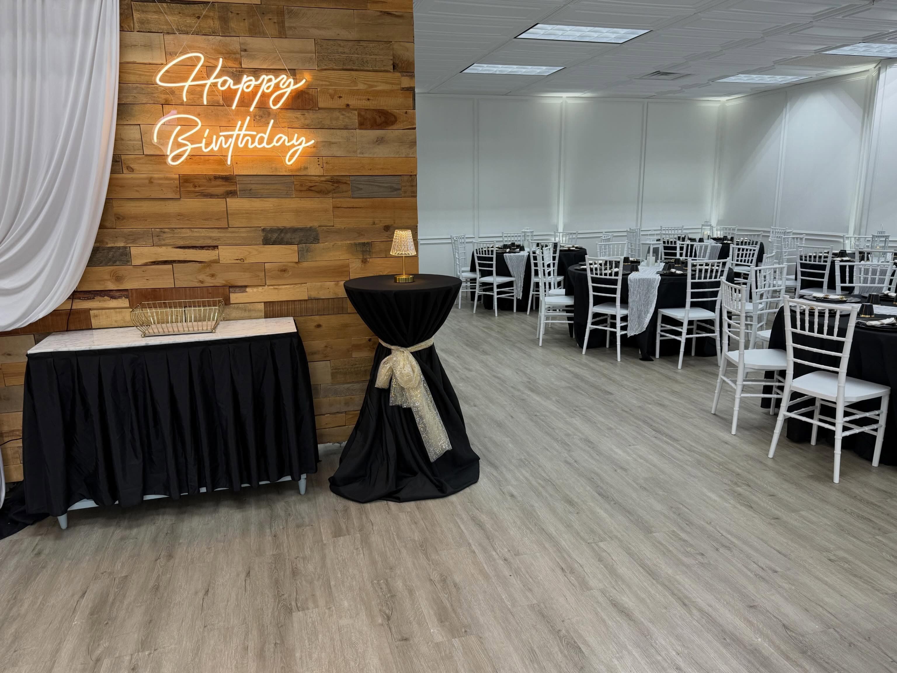 The Ehnita Suite dressed for a party with round tables, black linens, white chairs, and a wood accent wall lit by a neon sign