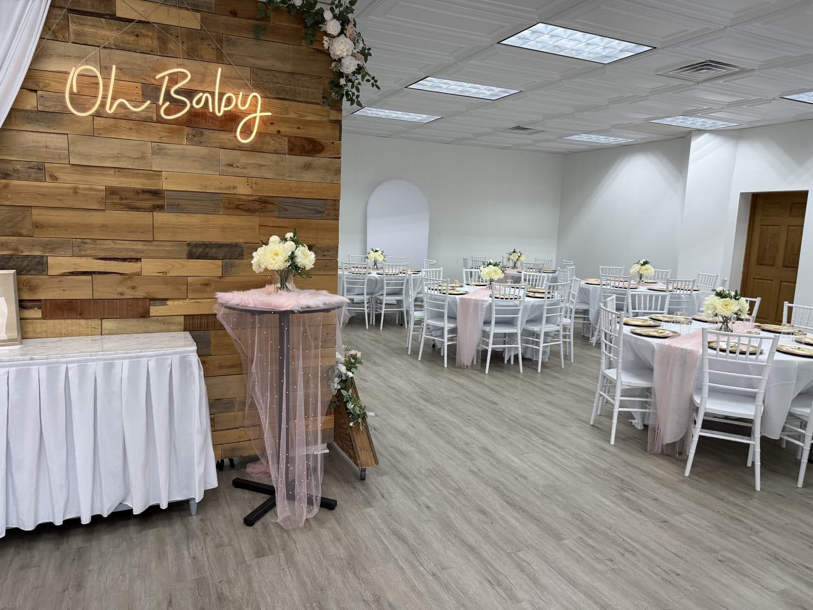 The Oh Baby neon sign glowing on the wood accent wall over a shower setup with blush tables